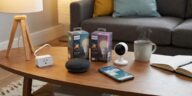 A collection of smart home devices including a Google Nest Mini, Philips Hue bulbs, a white security camera, and a smart plug on a coffee table next to a smartphone showing an automation app.