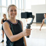A realistic shot of a blonde female athlete holding a smartphone on a stabilizer gimbal to record a vlog in a bright indoor gym, representing how female TikTok athletes 2025 are dominating sports media through content creation.