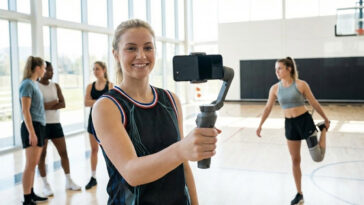 A realistic shot of a blonde female athlete holding a smartphone on a stabilizer gimbal to record a vlog in a bright indoor gym, representing how female TikTok athletes 2025 are dominating sports media through content creation.