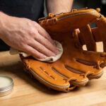 A close-up of hands rubbing leather conditioner into the pocket of a tan baseball glove on a wooden workbench, with a tin of conditioner and a baseball nearby.