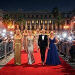 A realistic high-definition night scene of a red carpet event featuring celebrities in elegant 2025 fashion gowns walking past a blurred background of paparazzi and flashing cameras.