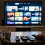 A first-person view of a person holding a remote control in a dimly lit living room, with several other remotes on a coffee table and a large smart TV displaying a streaming service interface in the background.