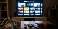 A first-person view of a person holding a remote control in a dimly lit living room, with several other remotes on a coffee table and a large smart TV displaying a streaming service interface in the background.